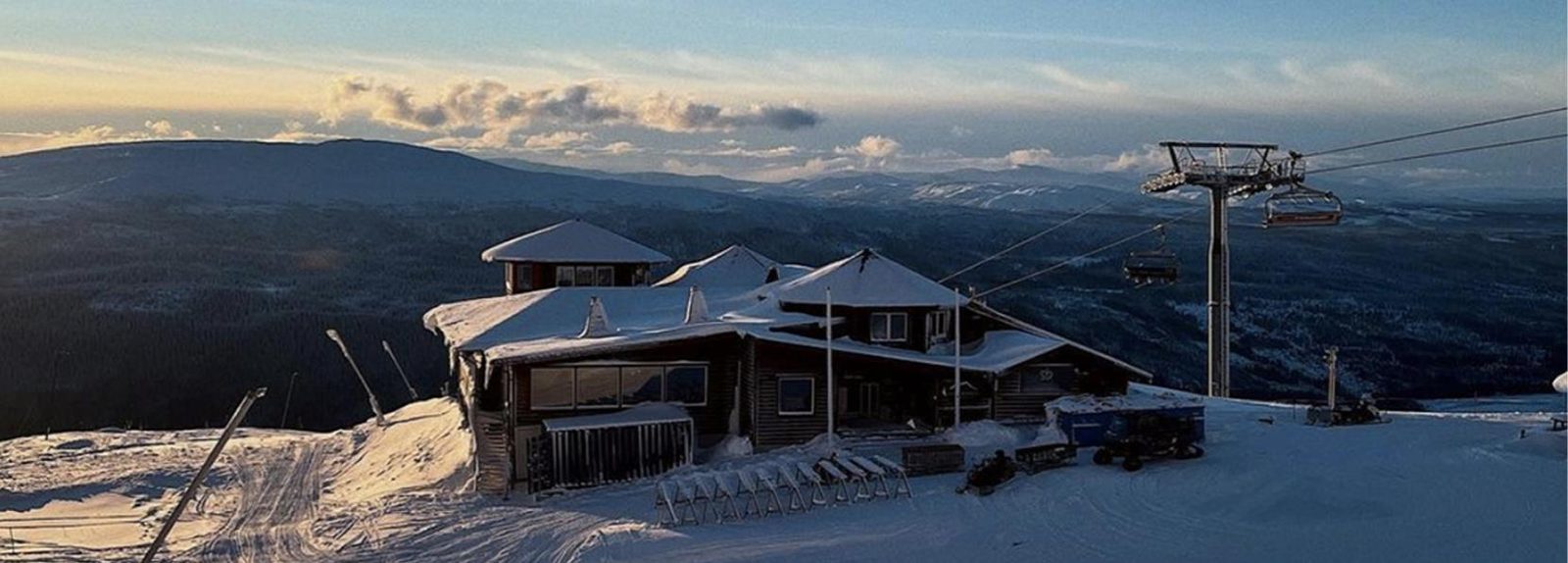 Restaurangbyggnad på snötäckt fjälltopp i Åre i kvällsljus, med liftlinje och vidsträckt fjällandskap bakom.